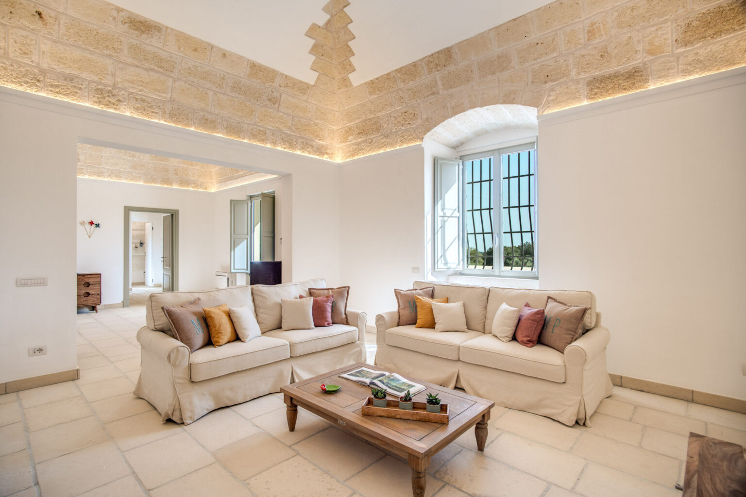 Bright and spacious living room featuring cream-colored sofas with decorative pillows, a wooden coffee table, and large windows that allow natural light to fill the room, highlighting the stone wall and ceiling design. | Only on SopranoVillas