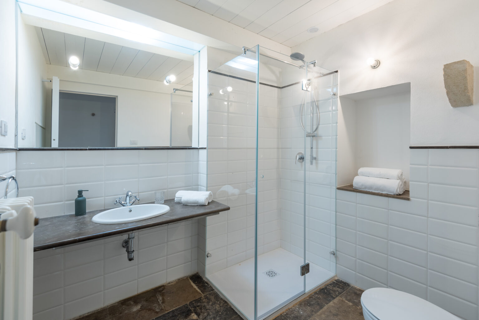 Modern bathroom interior featuring a glass shower enclosure, a sleek sink with a dark countertop, neatly stacked white towels, and stylish lighting. The space is designed with white tiled walls and a natural stone floor, creating a clean and inviting atmosphere. | Only on SopranoVillas