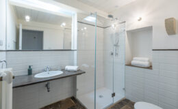 Modern bathroom interior featuring a glass shower enclosure, a sleek sink with a dark countertop, neatly stacked white towels, and stylish lighting. The space is designed with white tiled walls and a natural stone floor, creating a clean and inviting atmosphere.