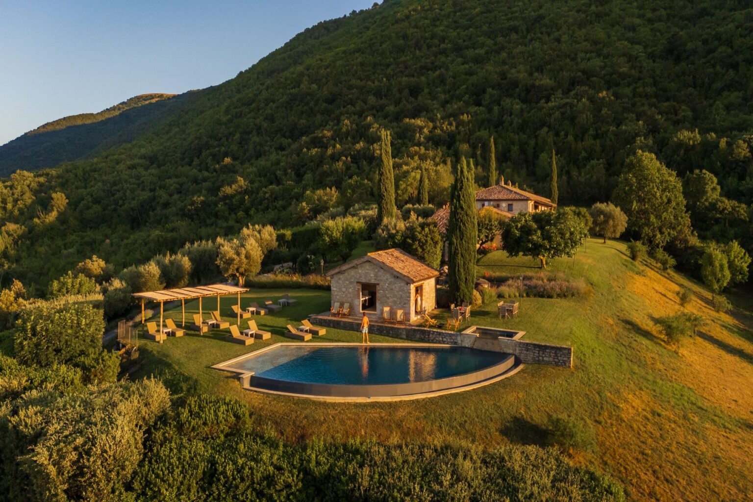 Aerial view of a luxurious countryside villa with a swimming pool, surrounded by lush green hills and olive trees. The property features a stone house, sun loungers, and a pergola, creating a serene outdoor retreat ideal for relaxation and leisure. | Only on SopranoVillas