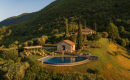 Aerial view of a luxurious countryside villa with a swimming pool, surrounded by lush green hills and olive trees. The property features a stone house, sun loungers, and a pergola, creating a serene outdoor retreat ideal for relaxation and leisure.