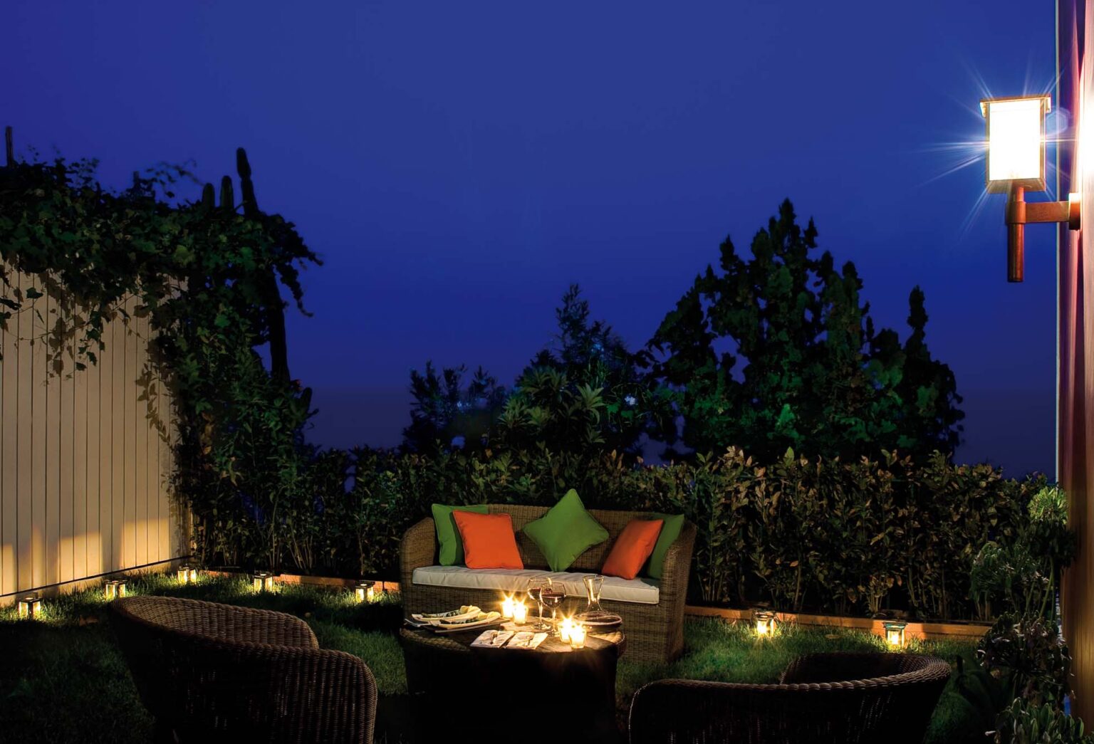 Cozy outdoor seating area with a sofa adorned with colorful cushions, surrounded by lush greenery and softly glowing lanterns, creating a serene ambiance at dusk. | Only on SopranoVillas