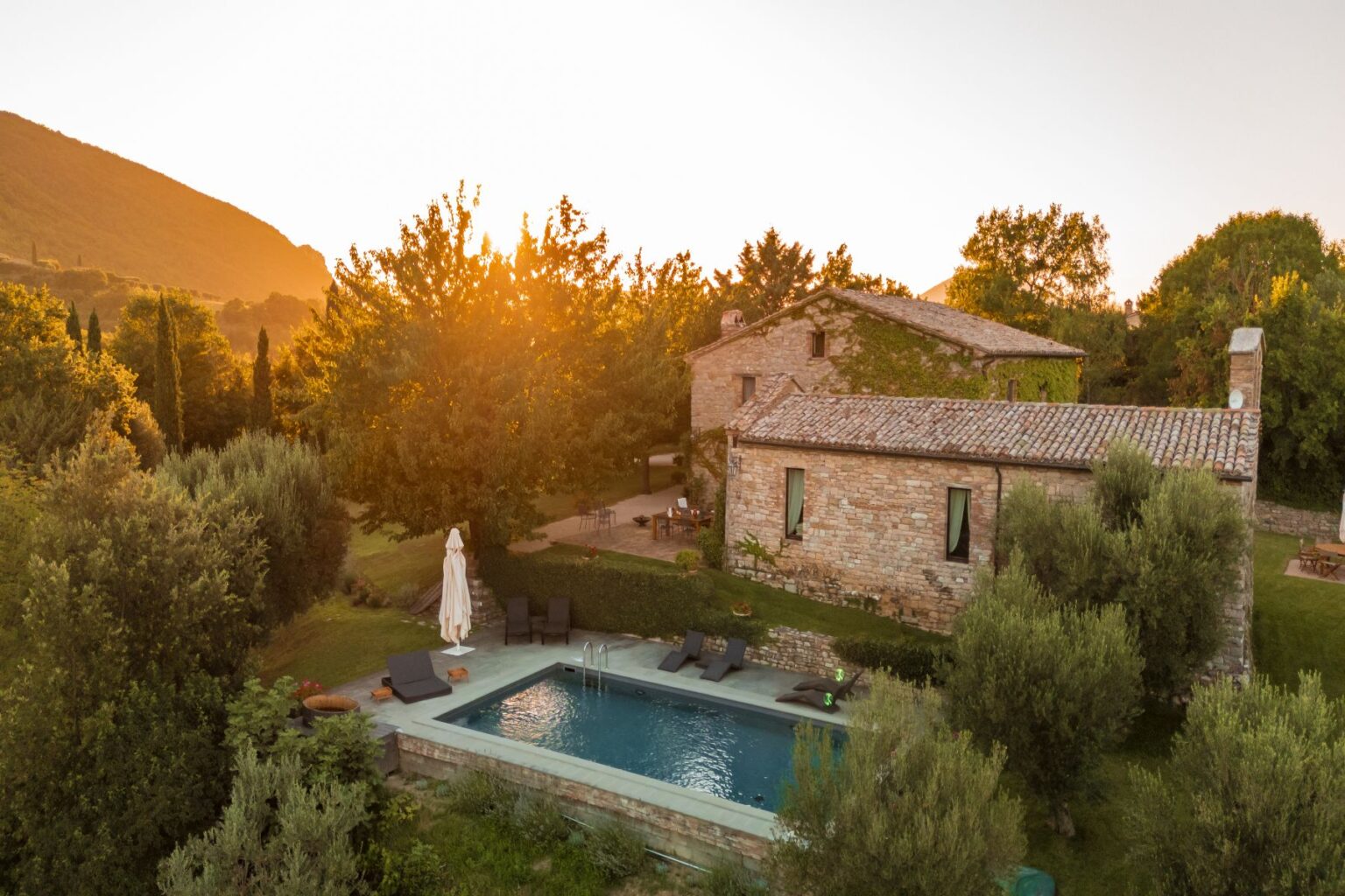 Aerial view of a rustic stone villa surrounded by lush greenery and trees, featuring a swimming pool and sun loungers, with a sunset illuminating the scene. Ideal for vacation rentals or countryside retreats. | Only on SopranoVillas