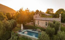 Aerial view of a rustic stone villa surrounded by lush greenery and trees, featuring a swimming pool and sun loungers, with a sunset illuminating the scene. Ideal for vacation rentals or countryside retreats.