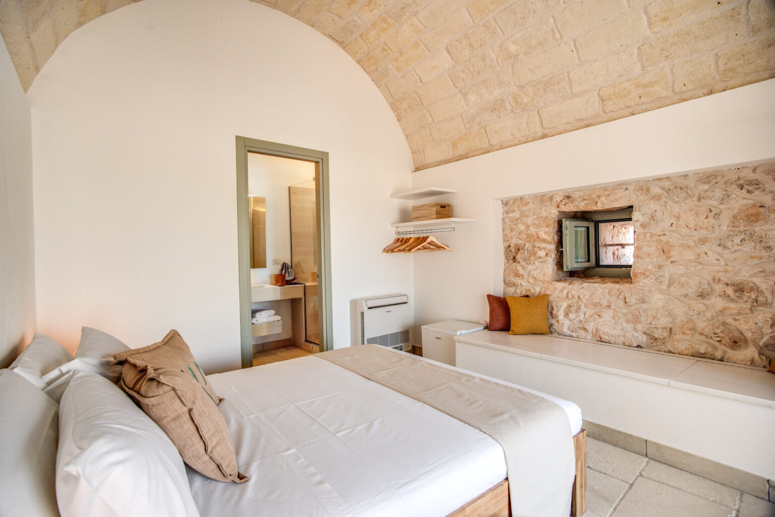Modern bedroom with a stone wall, arched ceiling, and minimalist decor. Features a comfortable bed with neutral-colored linens, a cozy seating area, and a small vanity with a mirror. Ideal for a relaxing getaway. | Only on SopranoVillas
