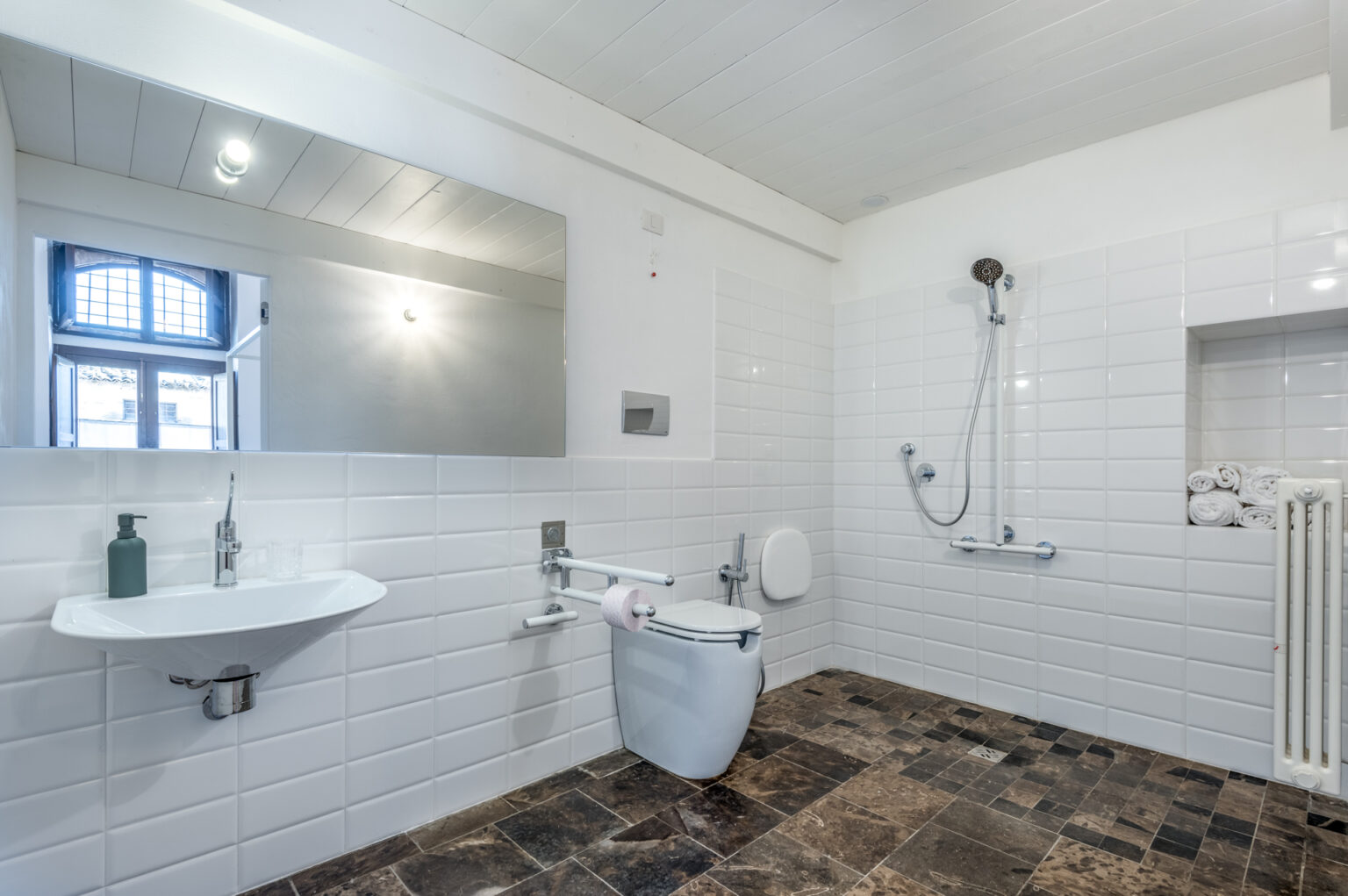 Modern bathroom featuring a sleek wall-mounted sink, a toilet with grab bars, a shower area with a handheld showerhead, and decorative towels. The space is bright with natural light from a window, complemented by white tiled walls and dark stone flooring. Ideal for accessibility and contemporary design. | Only on SopranoVillas