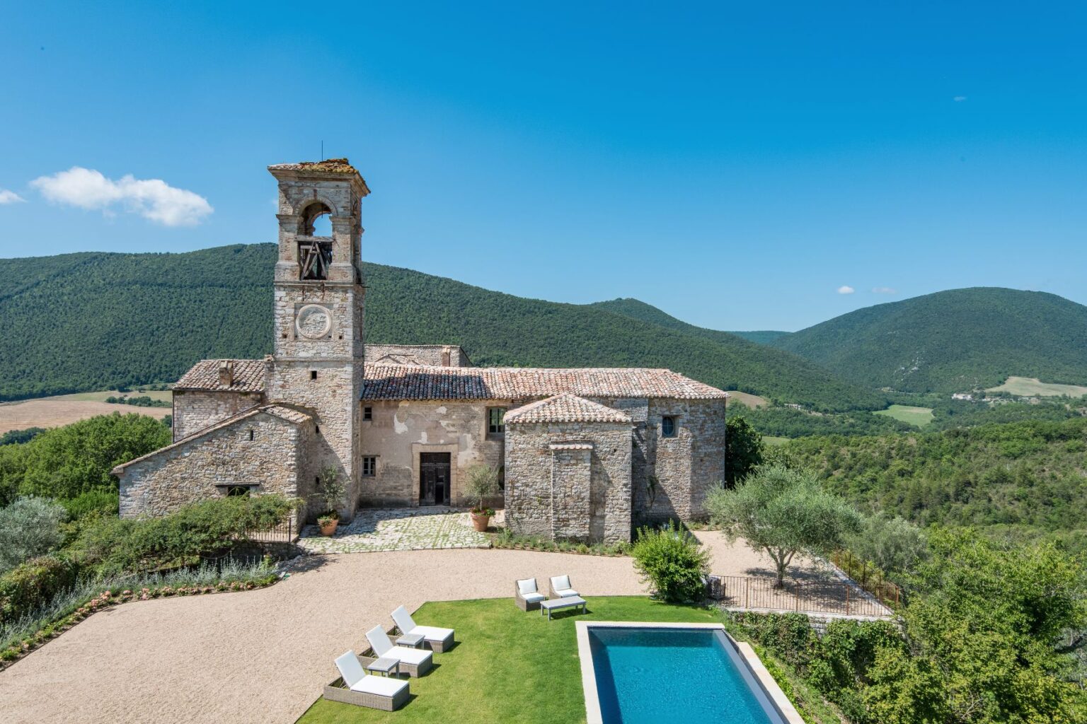 Historic stone villa with a bell tower, surrounded by lush green hills and a clear blue sky, featuring a swimming pool and sun loungers in the foreground. Ideal for luxury retreats and scenic getaways. | Only on SopranoVillas