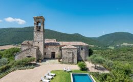Historic stone villa with a bell tower, surrounded by lush green hills and a clear blue sky, featuring a swimming pool and sun loungers in the foreground. Ideal for luxury retreats and scenic getaways.