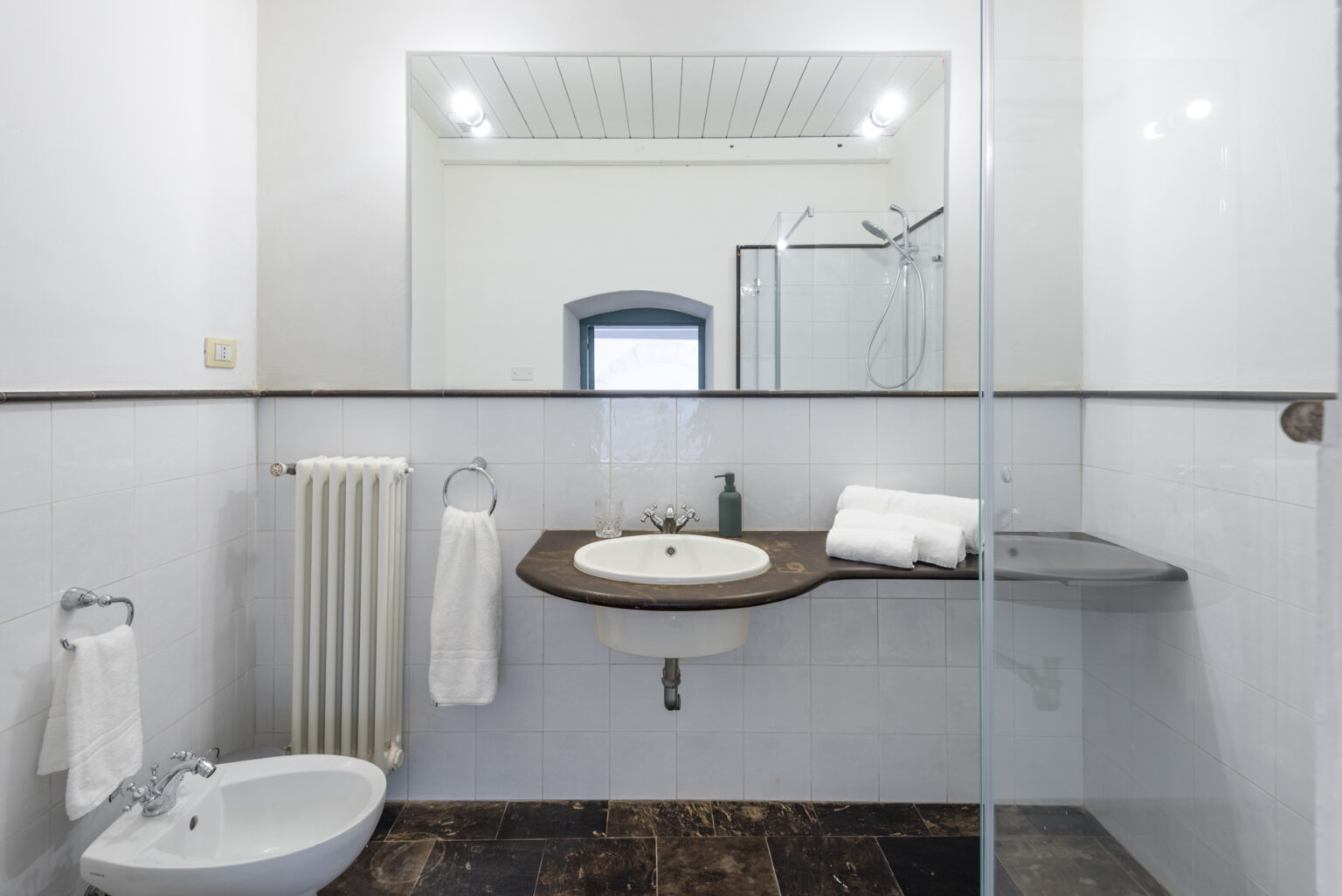 Modern bathroom interior featuring a bidet, sink with wooden countertop, towels, and a glass shower enclosure. The design emphasizes simplicity and functionality with neutral colors and ample natural light. | Only on SopranoVillas