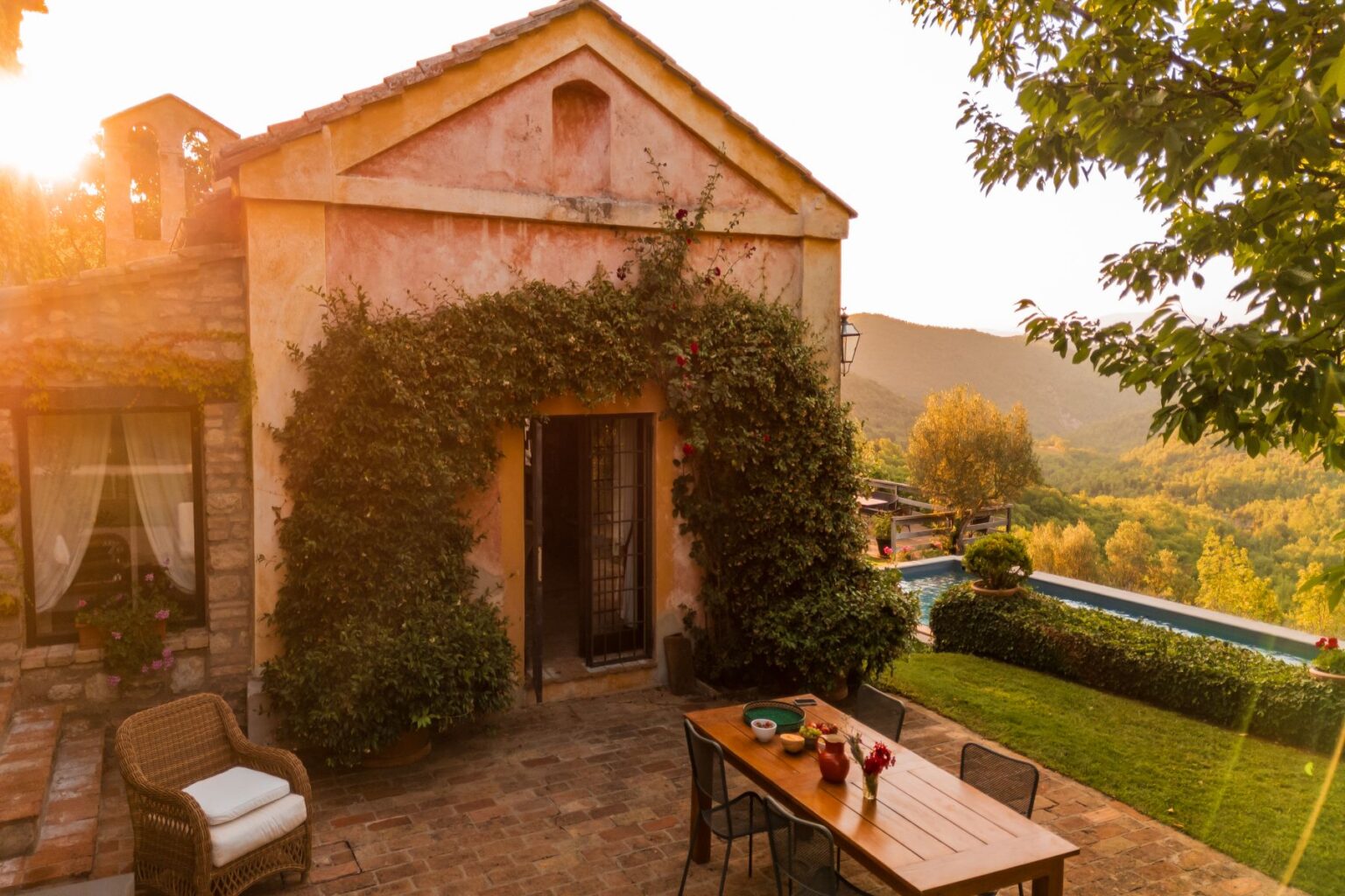 Charming rustic villa surrounded by lush greenery, featuring a wooden dining table set outdoors and a scenic view of rolling hills in the background at sunset. | Only on SopranoVillas