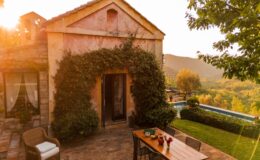 Charming rustic villa surrounded by lush greenery, featuring a wooden dining table set outdoors and a scenic view of rolling hills in the background at sunset.