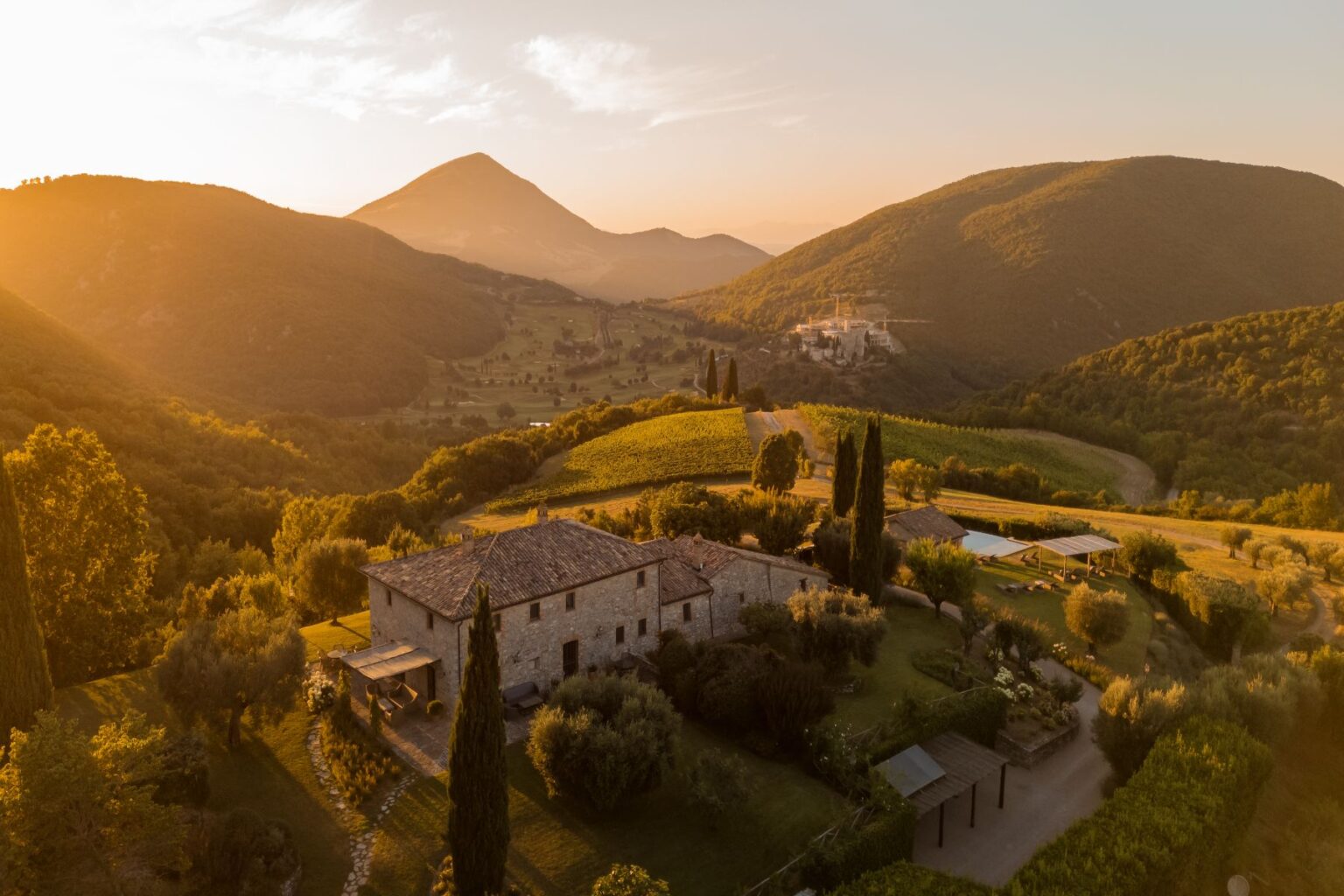 Scenic view of a rustic stone farmhouse surrounded by lush green hills and vineyards at sunset, highlighting the beauty of the Italian countryside with golden light illuminating the landscape. | Only on SopranoVillas