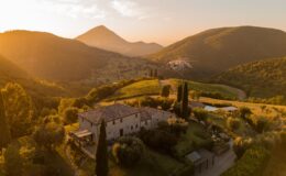 Scenic view of a rustic stone farmhouse surrounded by lush green hills and vineyards at sunset, highlighting the beauty of the Italian countryside with golden light illuminating the landscape.