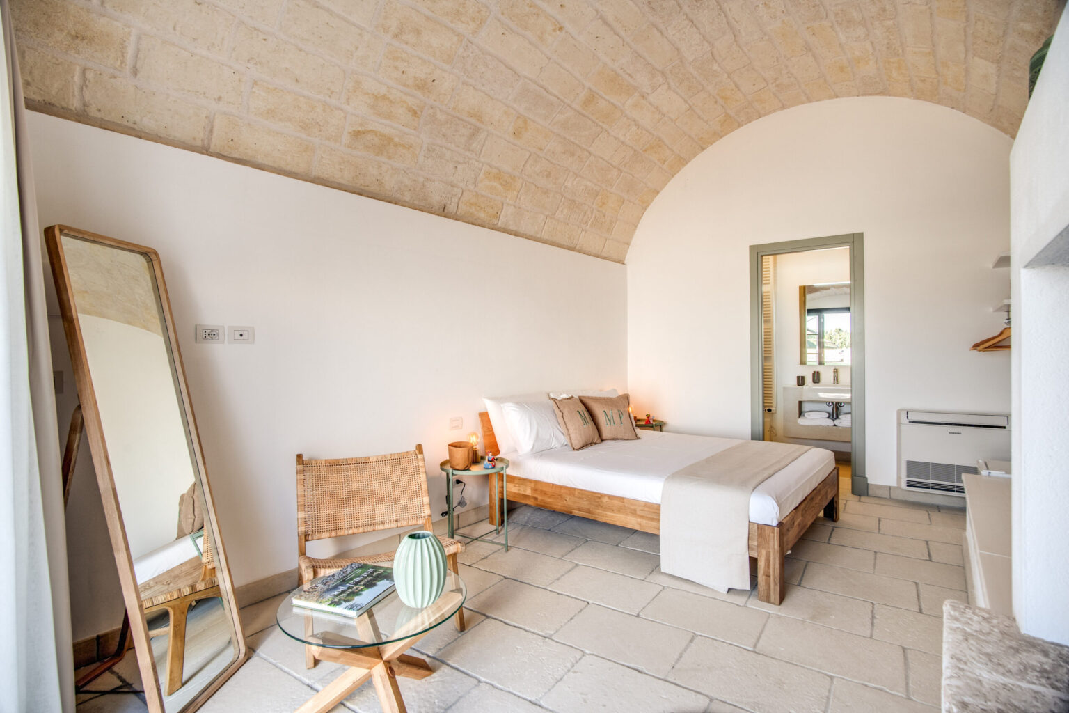Modern bedroom with a curved stone ceiling, featuring a wooden bed with neutral bedding, a large mirror, and a cozy seating area. Natural light floods the space, enhancing the minimalist decor and serene atmosphere. | Only on SopranoVillas
