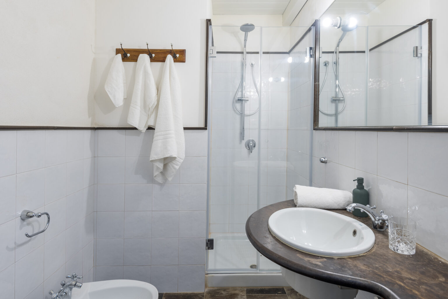 Modern bathroom featuring a glass shower enclosure, white towels hanging on a wooden rack, a round sink on a dark countertop, and a green soap dispenser. The clean design emphasizes functionality and style, ideal for a contemporary home. | Only on SopranoVillas