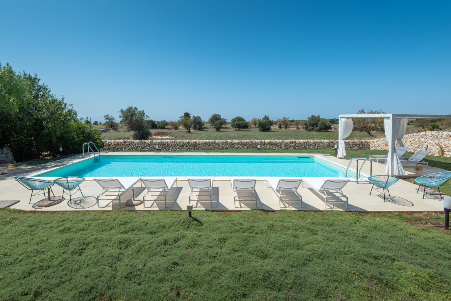 Modern outdoor swimming pool surrounded by lounge chairs and greenery, with a clear blue sky in the background, ideal for relaxation and leisure activities. | Only on SopranoVillas
