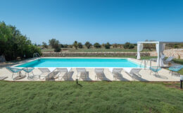 Modern outdoor swimming pool surrounded by lounge chairs and greenery, with a clear blue sky in the background, ideal for relaxation and leisure activities.