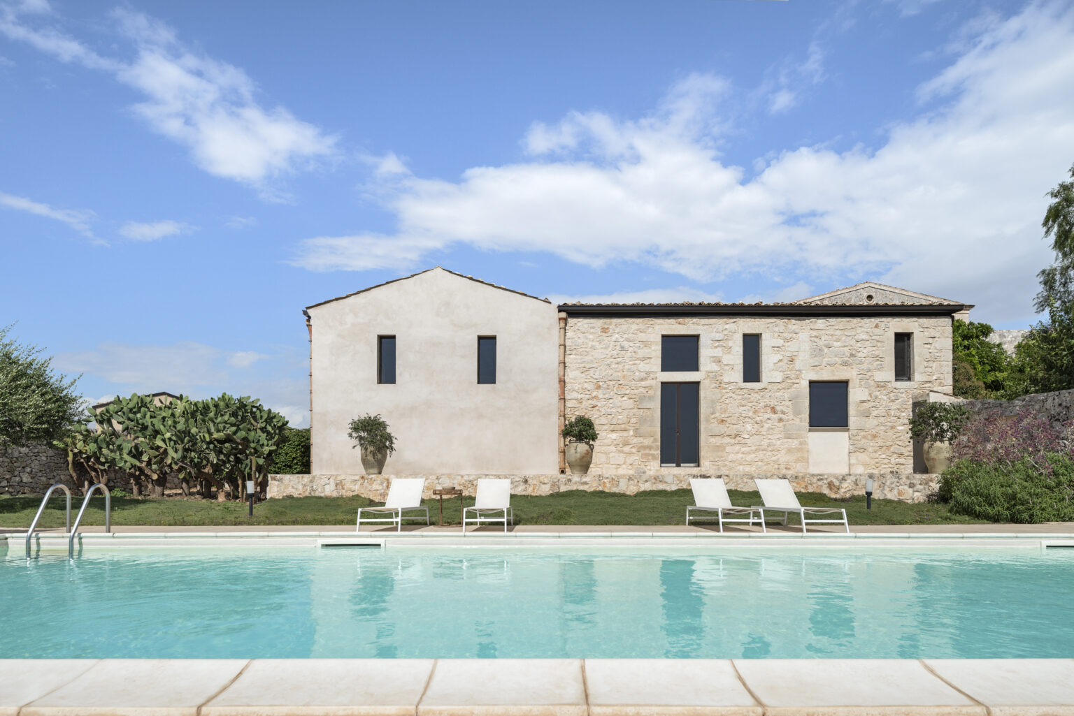 Modern stone house with large windows and a swimming pool in the foreground, surrounded by greenery and a clear blue sky. Ideal for luxury vacation rentals or architectural inspiration. | Only on SopranoVillas