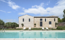 Modern stone house with large windows and a swimming pool in the foreground, surrounded by greenery and a clear blue sky. Ideal for luxury vacation rentals or architectural inspiration.