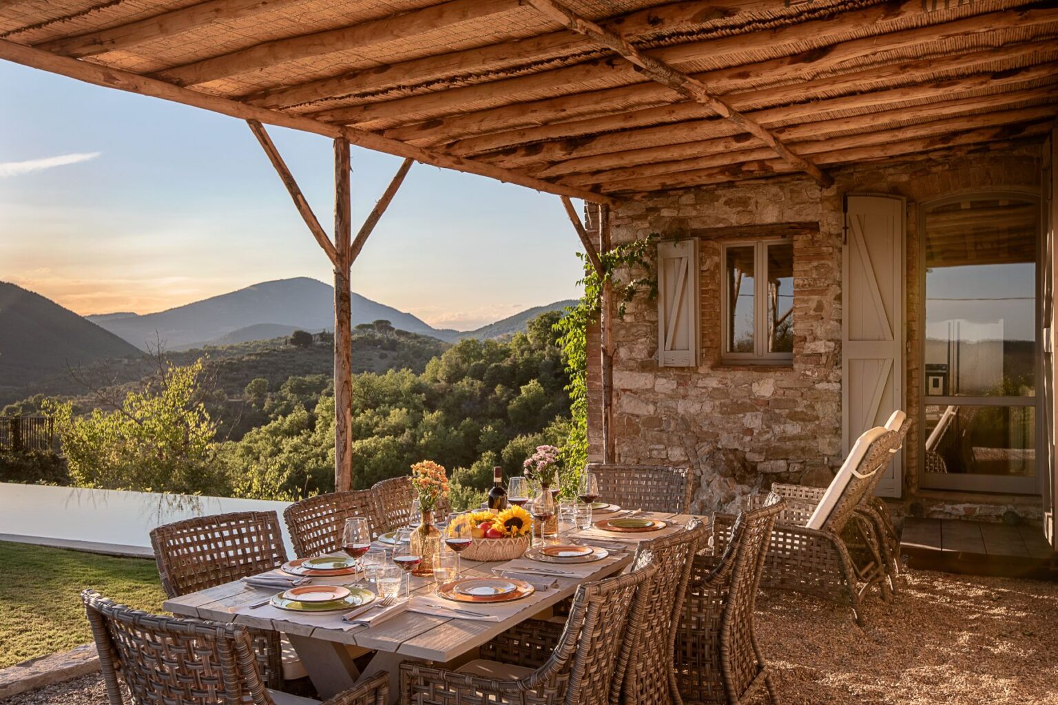 Outdoor dining area with a rustic wooden table set for a meal, featuring elegant tableware and glasses, surrounded by wicker chairs. The scene includes a stone house and a stunning view of rolling hills and mountains under a sunset sky, creating a tranquil atmosphere for al fresco dining. | Only on SopranoVillas