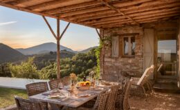 Outdoor dining area with a rustic wooden table set for a meal, featuring elegant tableware and glasses, surrounded by wicker chairs. The scene includes a stone house and a stunning view of rolling hills and mountains under a sunset sky, creating a tranquil atmosphere for al fresco dining.