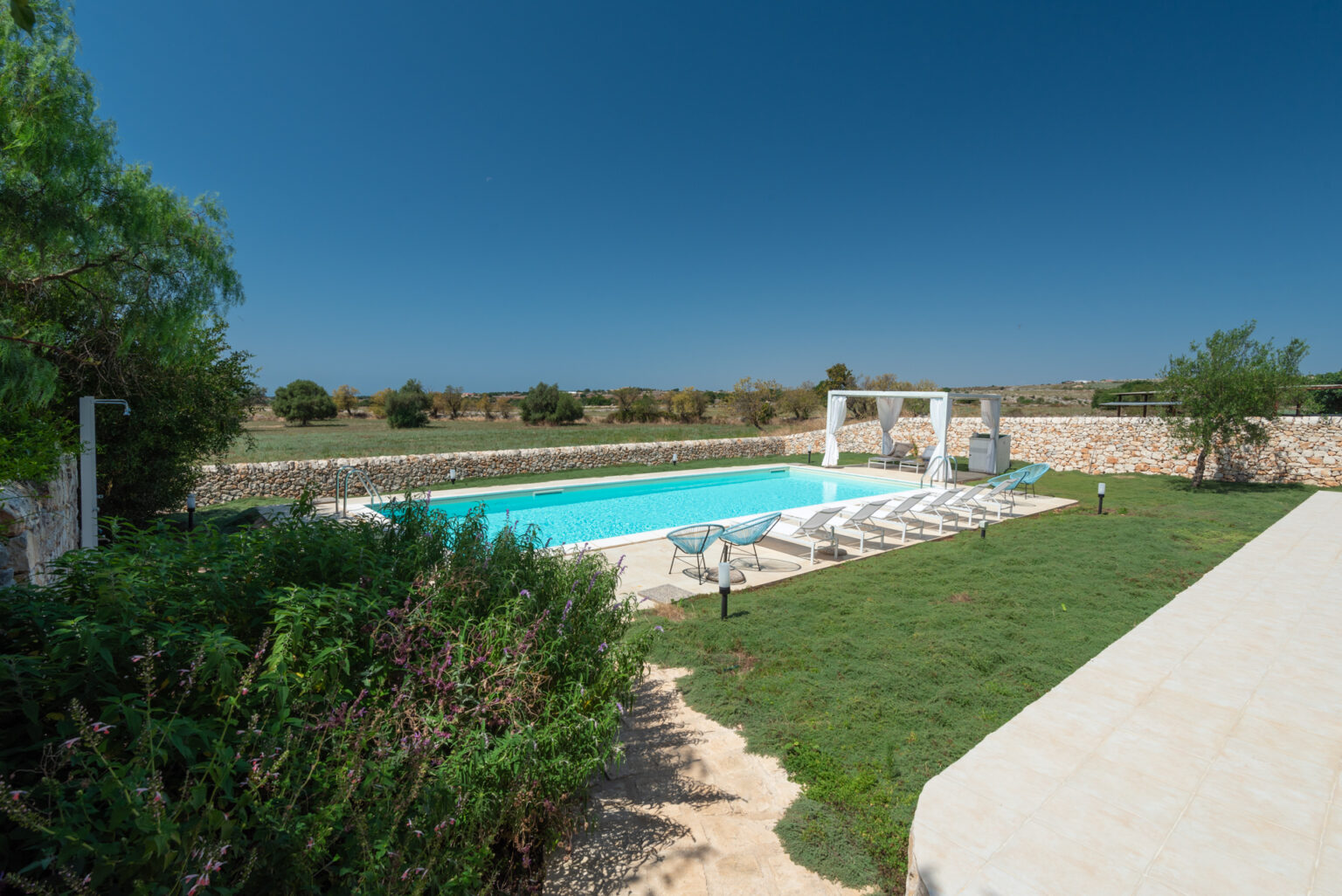Scenic view of a private swimming pool surrounded by lush greenery and stone walls, featuring sun loungers and a shaded seating area under a pergola, set against a clear blue sky. Ideal for relaxation and outdoor gatherings. | Only on SopranoVillas