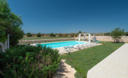 Scenic view of a private swimming pool surrounded by lush greenery and stone walls, featuring sun loungers and a shaded seating area under a pergola, set against a clear blue sky. Ideal for relaxation and outdoor gatherings.