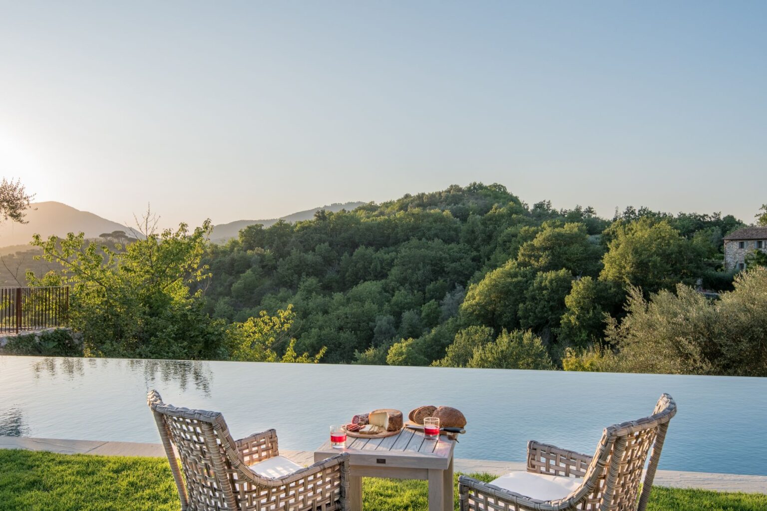 Serene outdoor setting with two wicker chairs facing an infinity pool, overlooking lush green hills and mountains at sunset, featuring a wooden table with snacks and drinks. Perfect for relaxation and scenic views. | Only on SopranoVillas