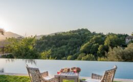 Serene outdoor setting with two wicker chairs facing an infinity pool, overlooking lush green hills and mountains at sunset, featuring a wooden table with snacks and drinks. Perfect for relaxation and scenic views.