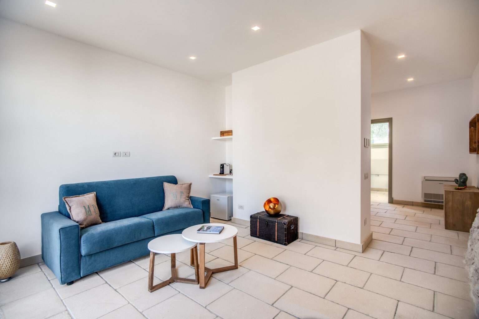Cozy living room featuring a blue sofa, two round coffee tables, and modern decor, with natural light streaming in through a window, showcasing a minimalist design and tiled floor. | Only on SopranoVillas