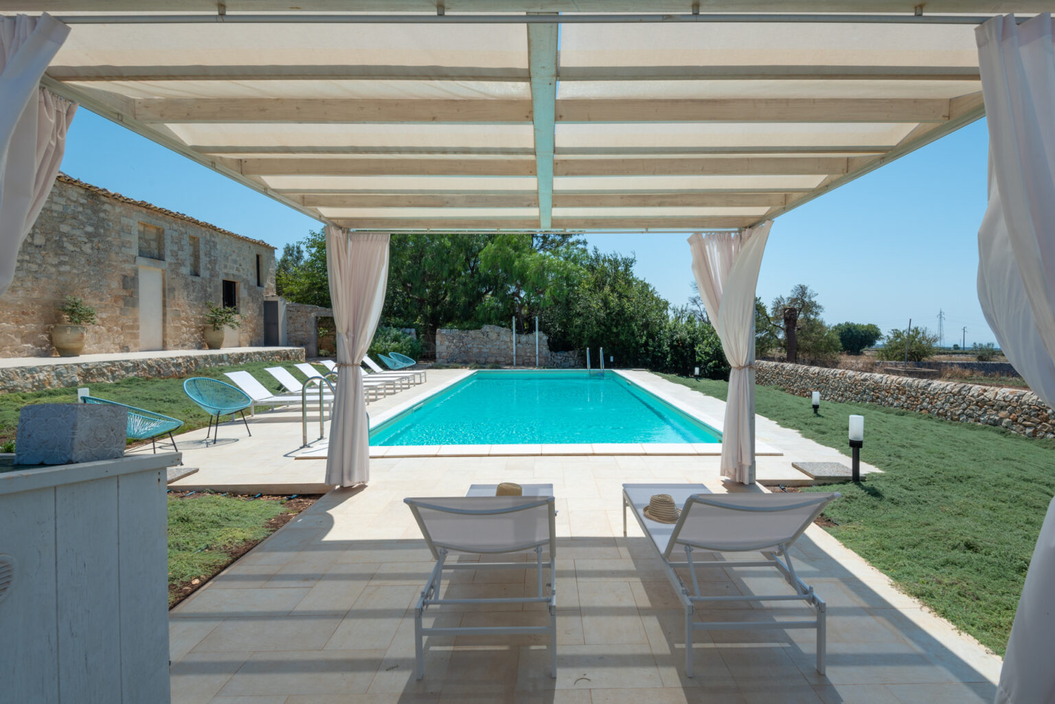 Luxurious outdoor area featuring a serene swimming pool surrounded by sun loungers and lush greenery, with a view of a rustic stone house under a shaded pergola. Ideal for relaxation and leisure in a tranquil setting. | Only on SopranoVillas