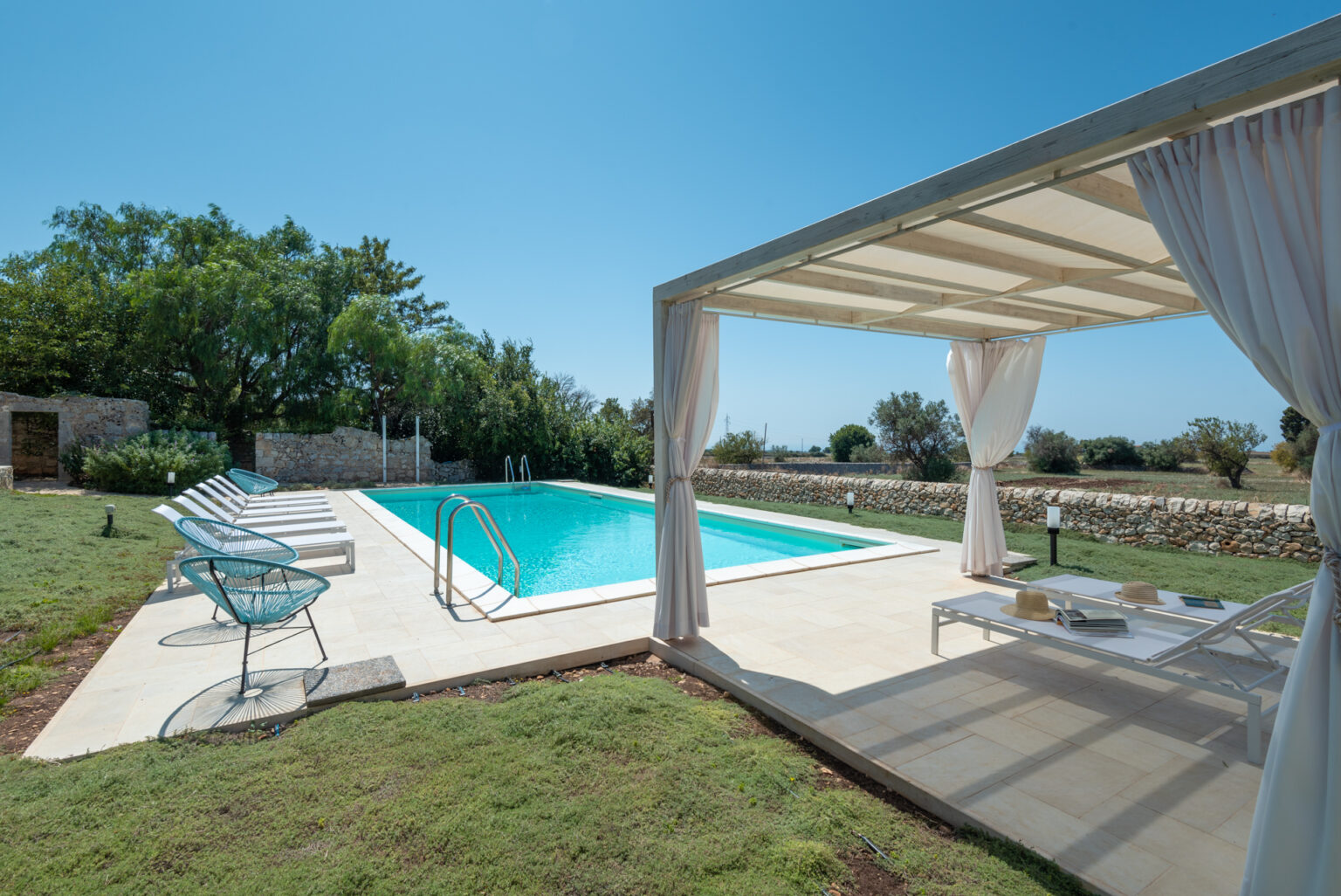 Modern outdoor pool area featuring a sparkling blue swimming pool, comfortable lounge chairs, and a shaded seating area with draped curtains, surrounded by lush greenery and stone walls, perfect for relaxation and leisure. | Only on SopranoVillas