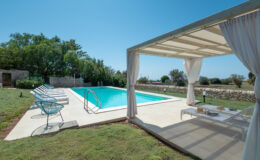 Modern outdoor pool area featuring a sparkling blue swimming pool, comfortable lounge chairs, and a shaded seating area with draped curtains, surrounded by lush greenery and stone walls, perfect for relaxation and leisure.
