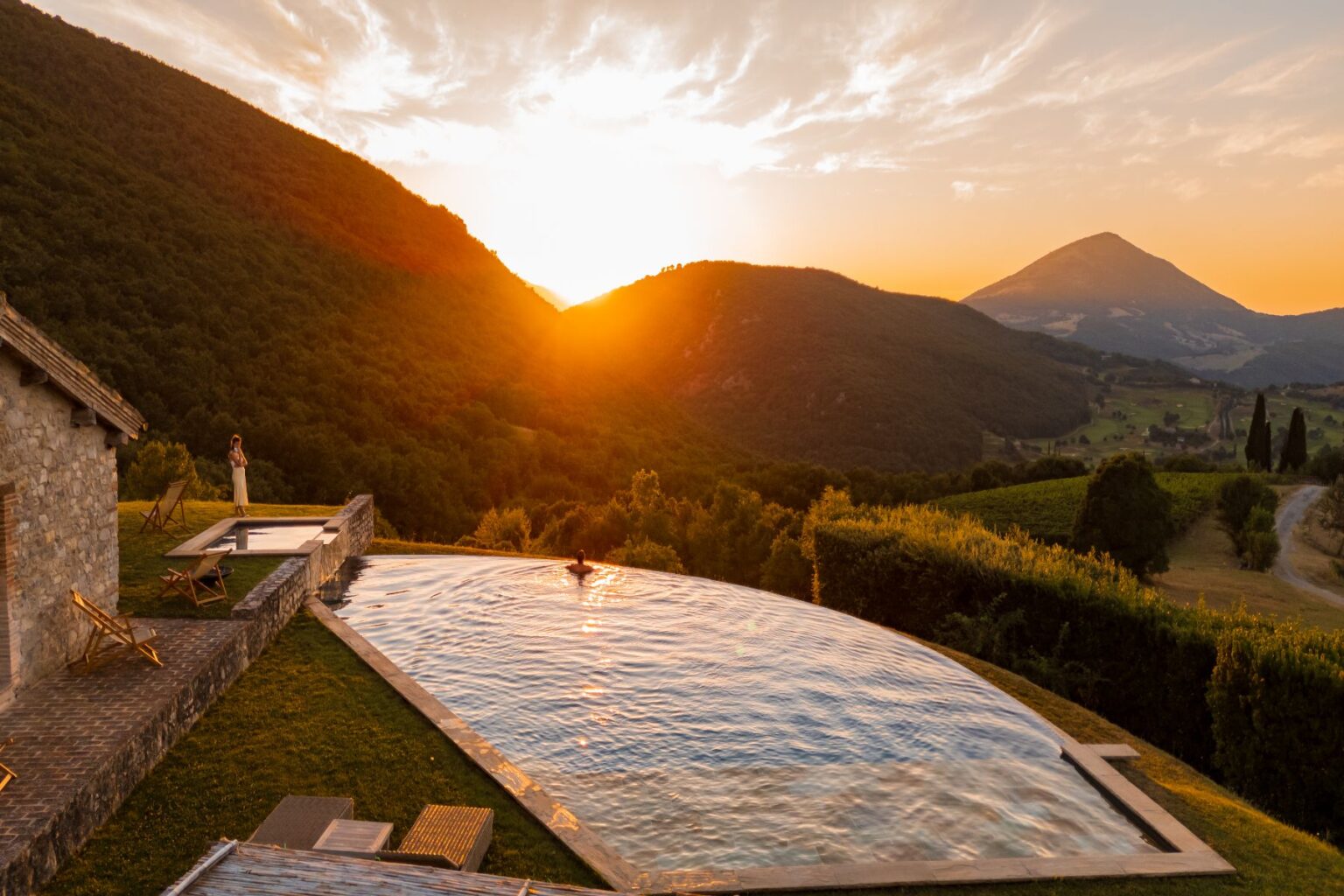 Stunning sunset view over a luxury villa with an infinity pool, surrounded by lush green hills and mountains, creating a serene and picturesque landscape ideal for relaxation and nature lovers. | Only on SopranoVillas