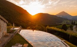 Stunning sunset view over a luxury villa with an infinity pool, surrounded by lush green hills and mountains, creating a serene and picturesque landscape ideal for relaxation and nature lovers.
