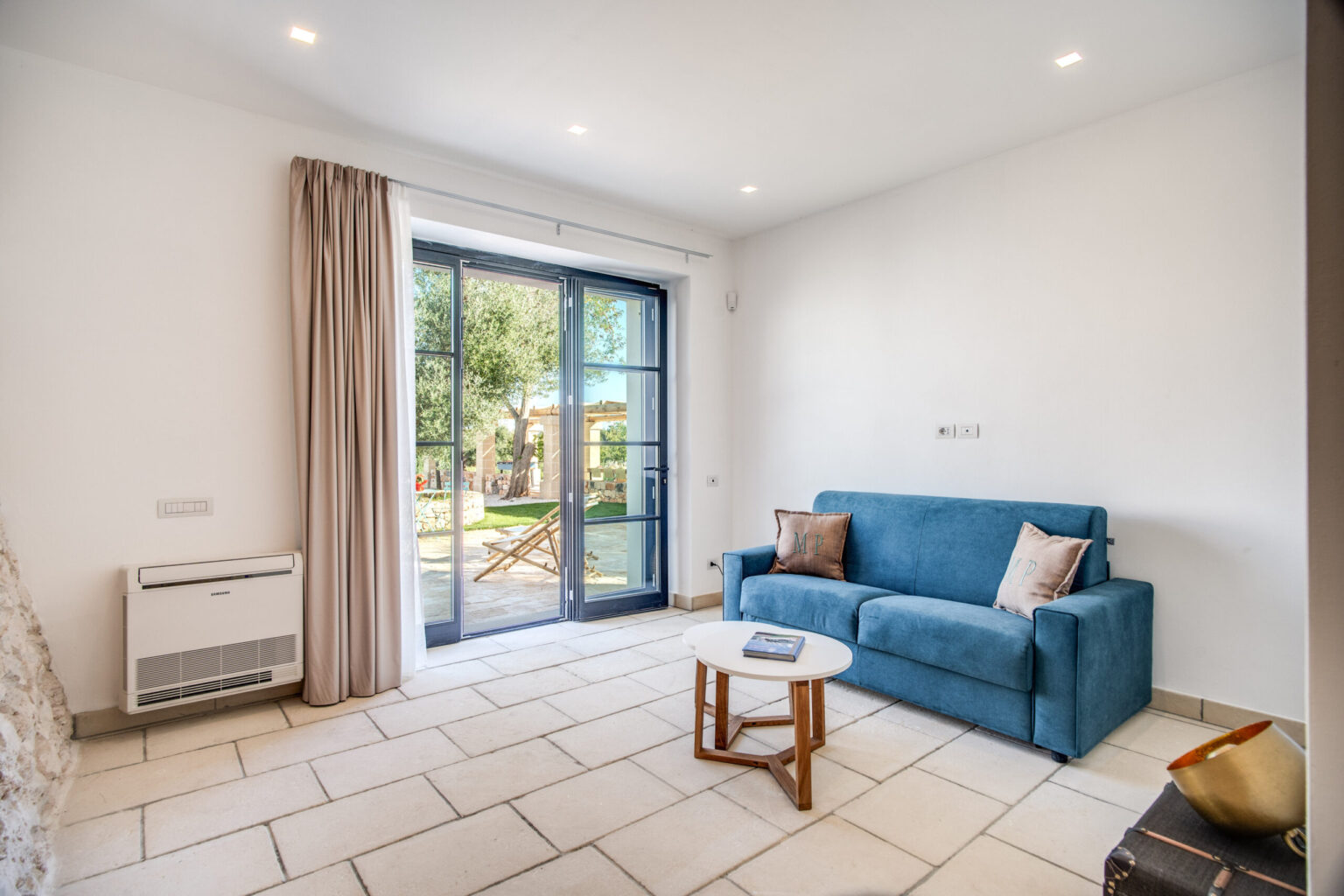 Modern living room featuring a blue sofa, a wooden coffee table, and large glass doors opening to a garden. The space is bright and airy, with neutral-colored walls and tiled flooring. | Only on SopranoVillas