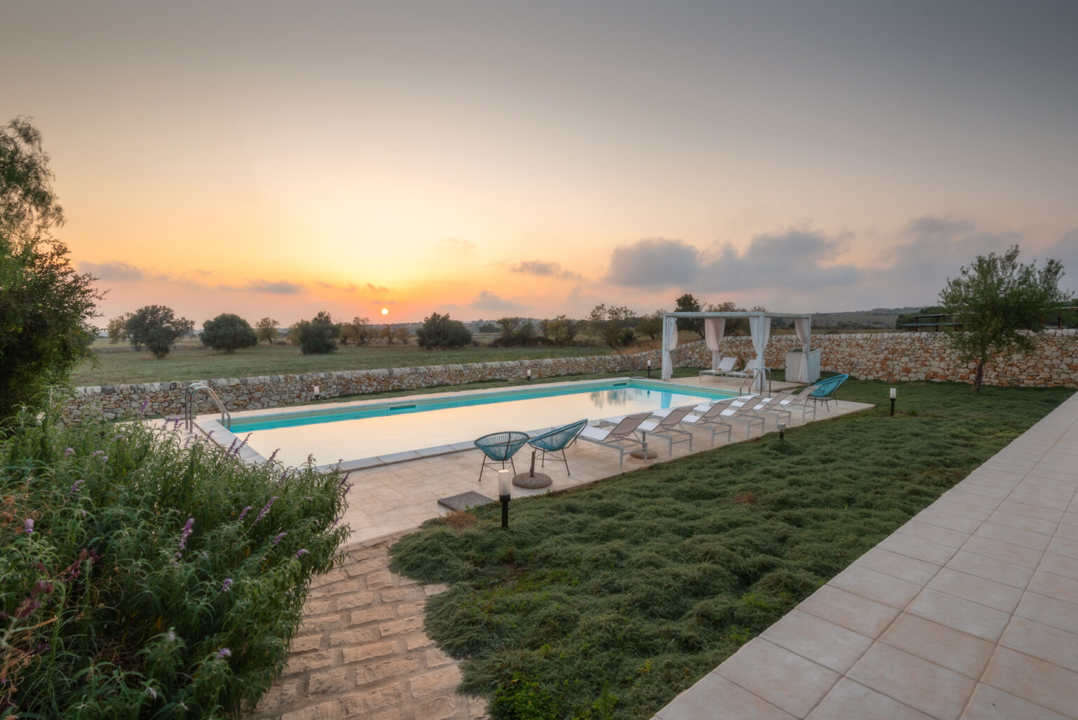 Serene outdoor pool area at sunset, featuring lounge chairs and a shaded seating area, surrounded by lush greenery and a stone wall, ideal for relaxation and leisure. | Only on SopranoVillas