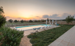 Serene outdoor pool area at sunset, featuring lounge chairs and a shaded seating area, surrounded by lush greenery and a stone wall, ideal for relaxation and leisure.