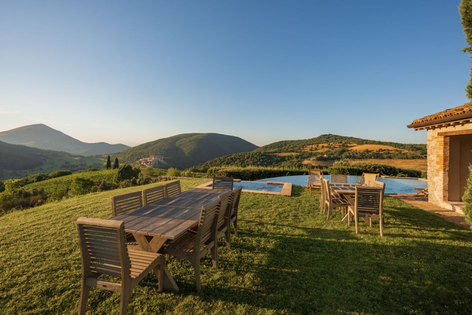 Outdoor dining area with wooden tables and chairs overlooking a scenic landscape of rolling hills and a swimming pool, ideal for relaxation and gatherings. | Only on SopranoVillas