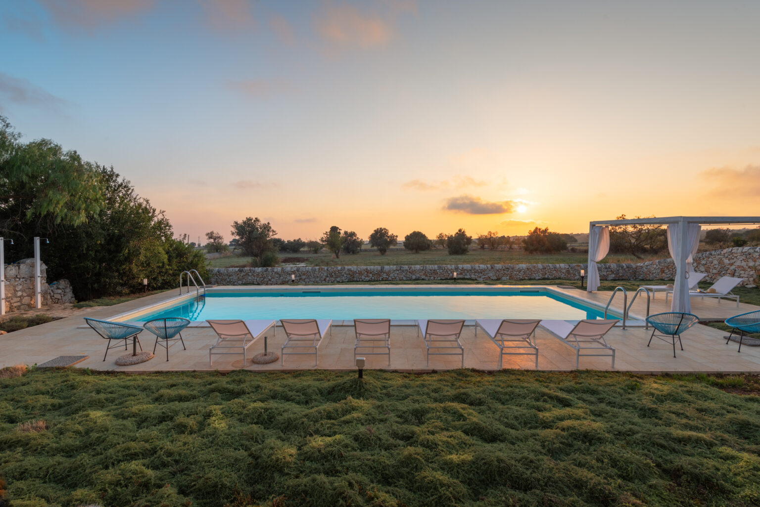 Serene outdoor swimming pool at sunset, surrounded by lush greenery and comfortable lounge chairs, perfect for relaxation and leisure. | Only on SopranoVillas