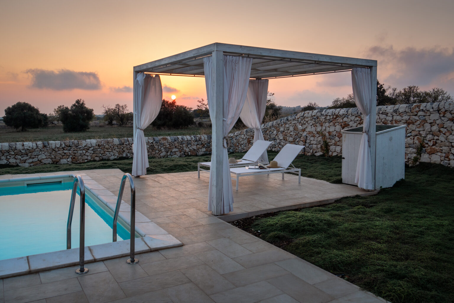 Serene outdoor pool area at sunset featuring a white pergola with draped curtains, two lounge chairs, and a stone wall backdrop, ideal for relaxation and leisure. | Only on SopranoVillas