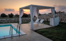 Serene outdoor pool area at sunset featuring a white pergola with draped curtains, two lounge chairs, and a stone wall backdrop, ideal for relaxation and leisure.