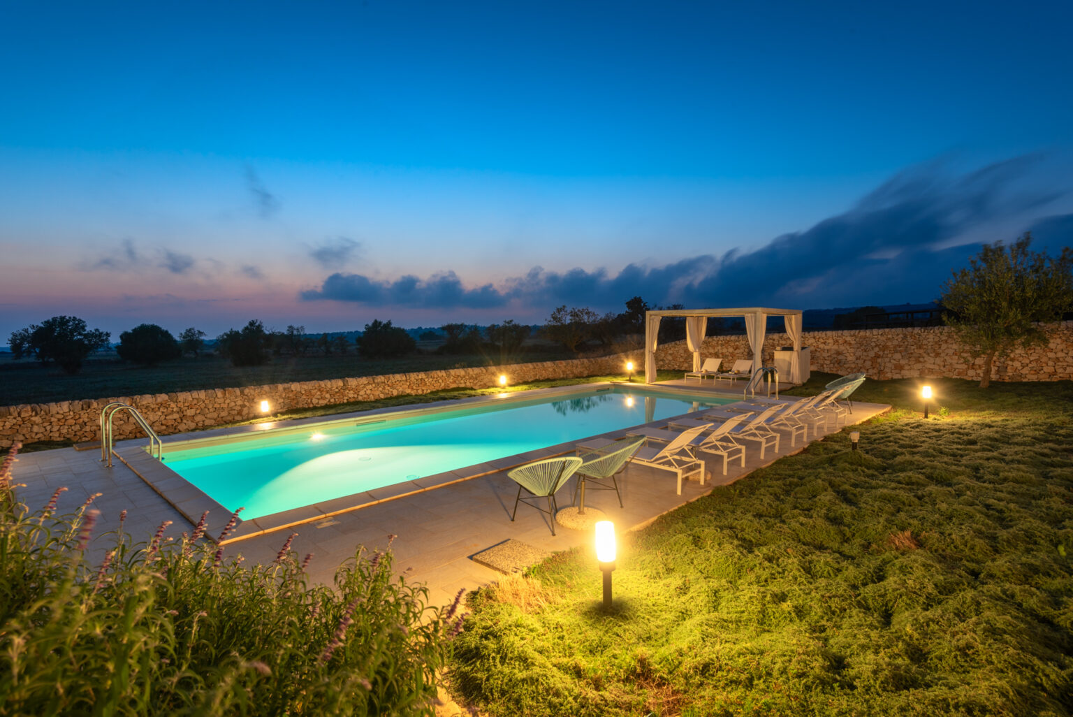 Luxurious outdoor swimming pool at dusk, surrounded by lush greenery and stone walls, featuring lounge chairs and a shaded seating area, creating a serene and inviting atmosphere for relaxation. | Only on SopranoVillas