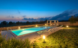 Luxurious outdoor swimming pool at dusk, surrounded by lush greenery and stone walls, featuring lounge chairs and a shaded seating area, creating a serene and inviting atmosphere for relaxation.