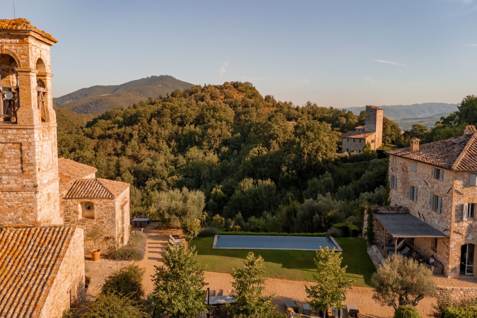 Aerial view of a rustic stone villa in the Italian countryside, featuring a bell tower, lush green hills, and a serene swimming pool surrounded by landscaped gardens. The scene captures the tranquility and natural beauty of the region, highlighting traditional architecture amidst scenic mountains. | Only on SopranoVillas