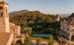 Aerial view of a rustic stone villa in the Italian countryside, featuring a bell tower, lush green hills, and a serene swimming pool surrounded by landscaped gardens. The scene captures the tranquility and natural beauty of the region, highlighting traditional architecture amidst scenic mountains.