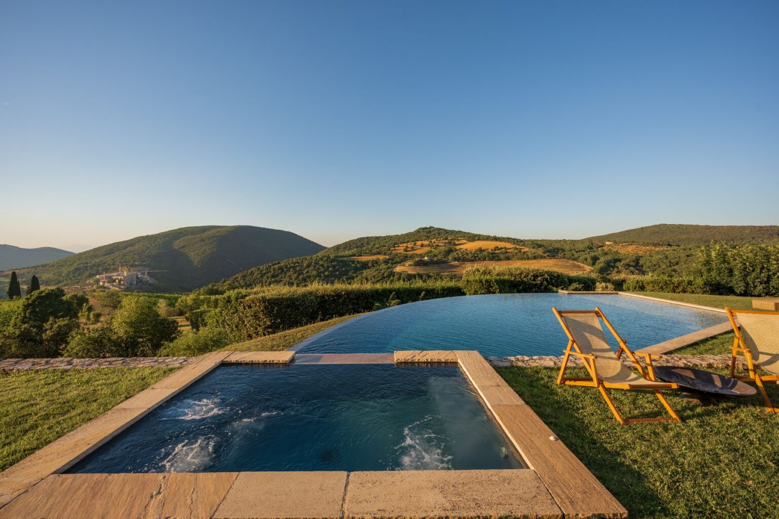 Scenic view of an infinity pool and a hot tub overlooking lush green hills and a clear blue sky, with wooden lounge chairs set on the grassy terrace, perfect for relaxation and enjoying nature. | Only on SopranoVillas