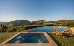 Scenic view of an infinity pool and a hot tub overlooking lush green hills and a clear blue sky, with wooden lounge chairs set on the grassy terrace, perfect for relaxation and enjoying nature.