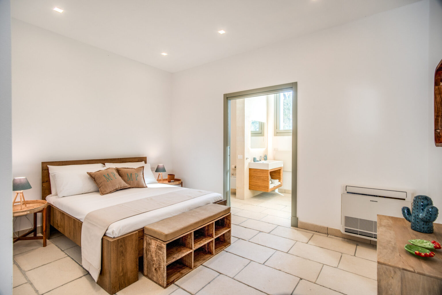 Cozy modern bedroom featuring a wooden bed with decorative pillows, a small side table with a lamp, and a bench with storage. Natural light floods in from an adjoining bathroom with a stylish sink and cabinetry. The room has a minimalist design with light-colored walls and tiled flooring, creating a serene atmosphere. | Only on SopranoVillas