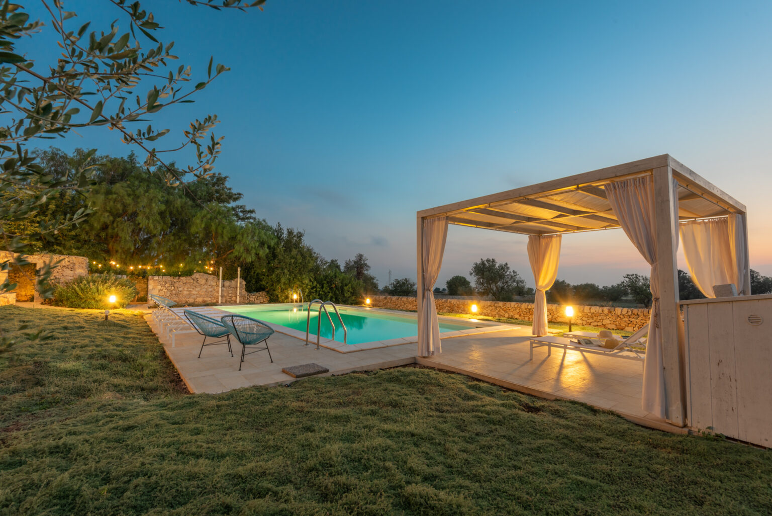 Serene outdoor pool area at dusk featuring a well-lit swimming pool, comfortable seating, and a shaded cabana, surrounded by lush greenery and stone walls, perfect for relaxation and gatherings. | Only on SopranoVillas